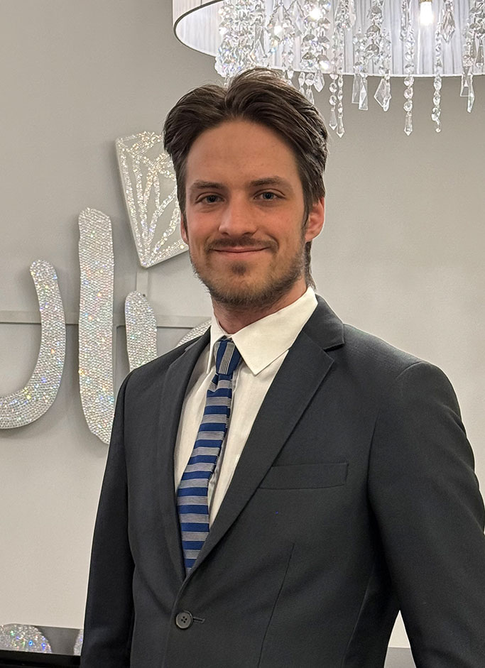 Ein Mann im Anzug lächelt; braune Haare, blau-gestreifte Krawatte, Kristallleuchter im Hintergrund.