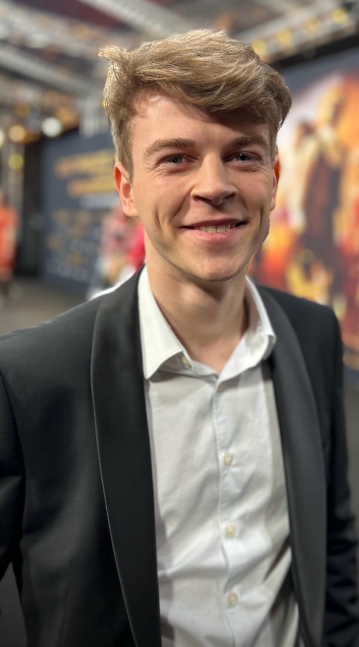 Ein lächelnder jüngerer Mann in Anzug und Hemd, braune Haare, bei einer Veranstaltung.