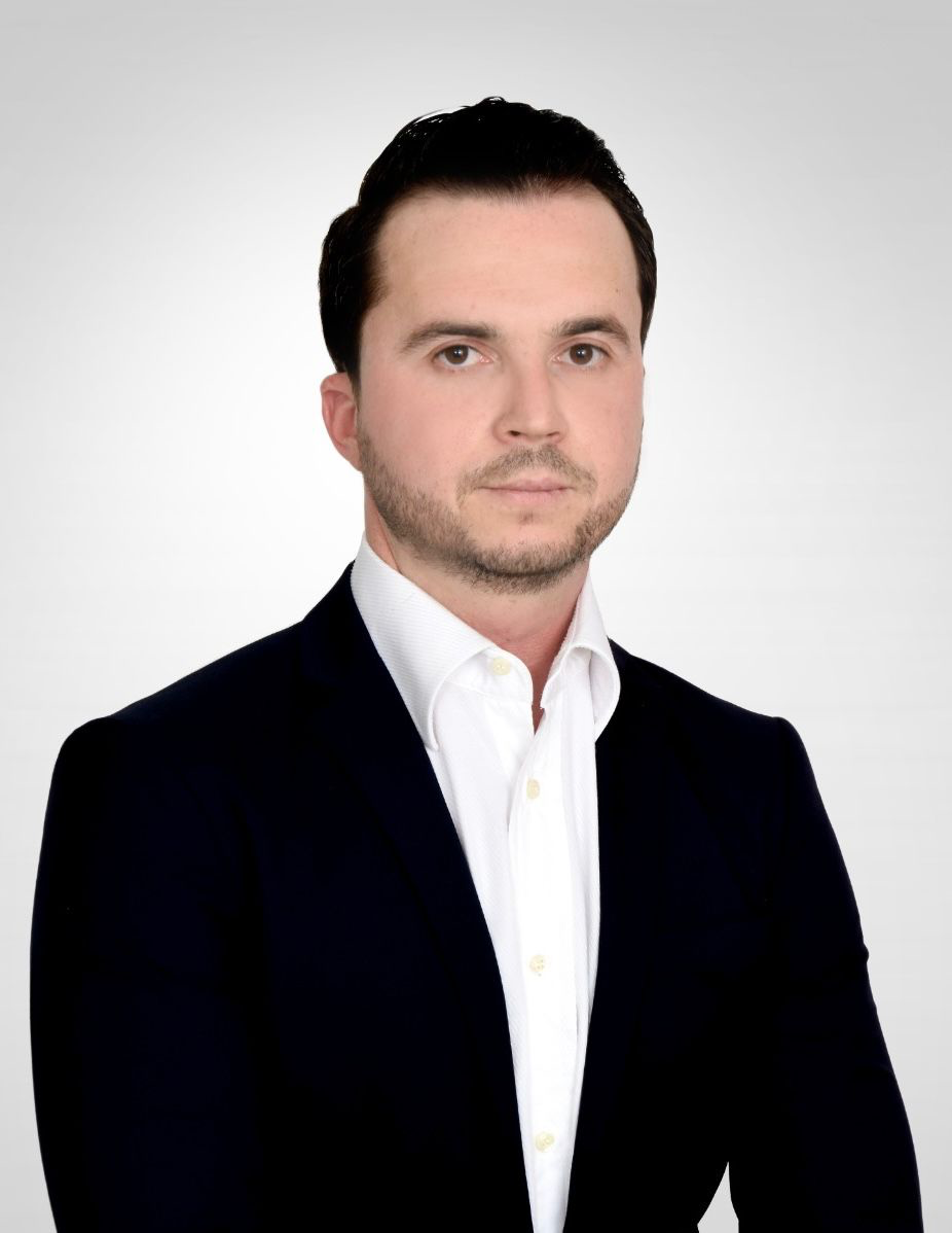Ein Mann im Anzug mit ernstem Gesichtsausdruck vor grauem Hintergrund.