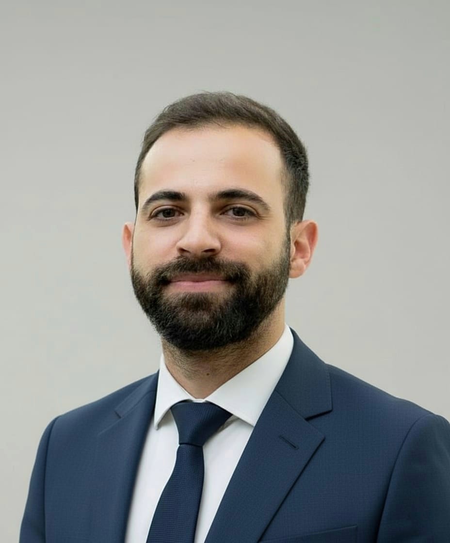 Ein lächelnder Mann mit dunklem Bart im blauen Anzug vor grauem Hintergrund